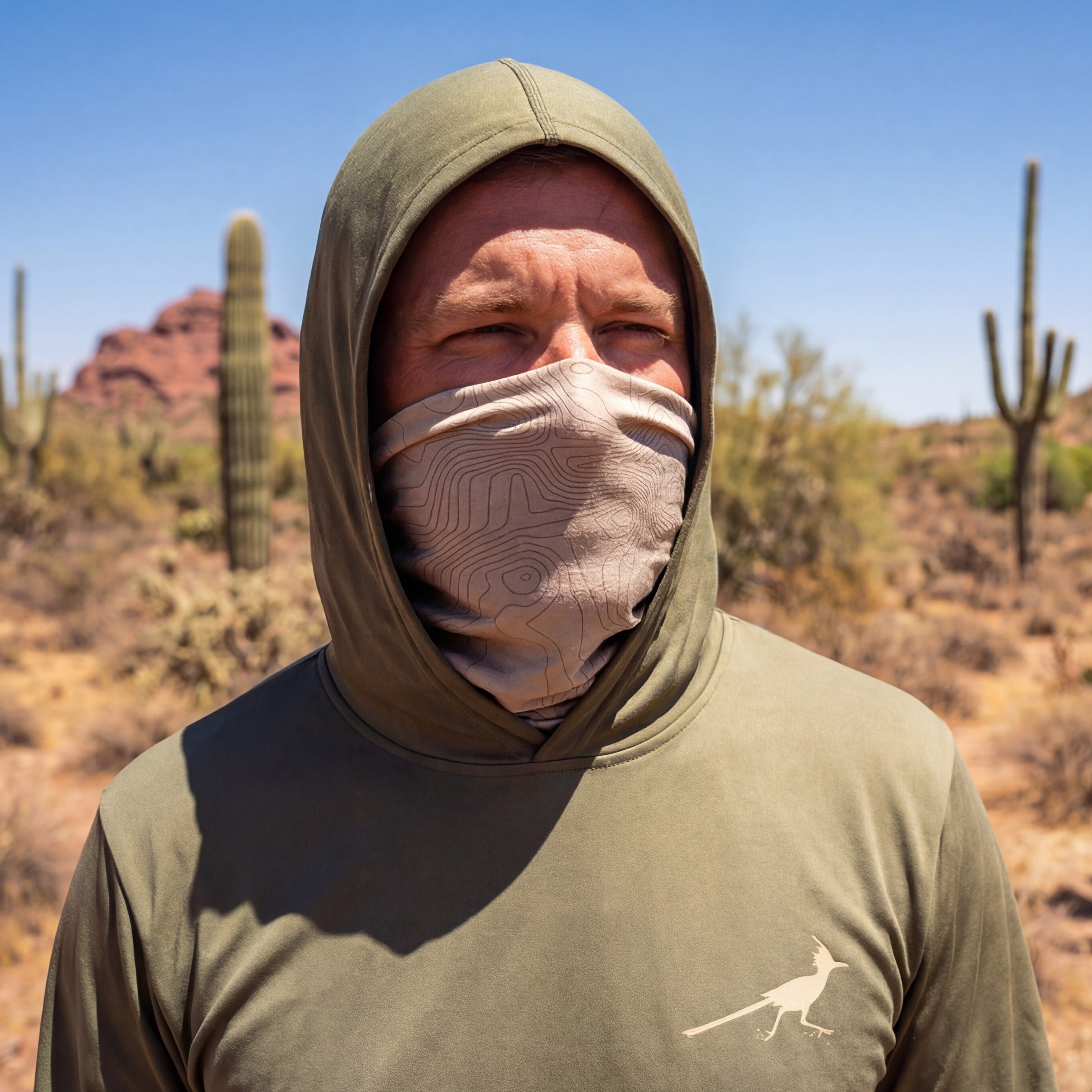 Man wearing a green hooded UPF shirt and neck gaiter in a desert landscape with cacti.