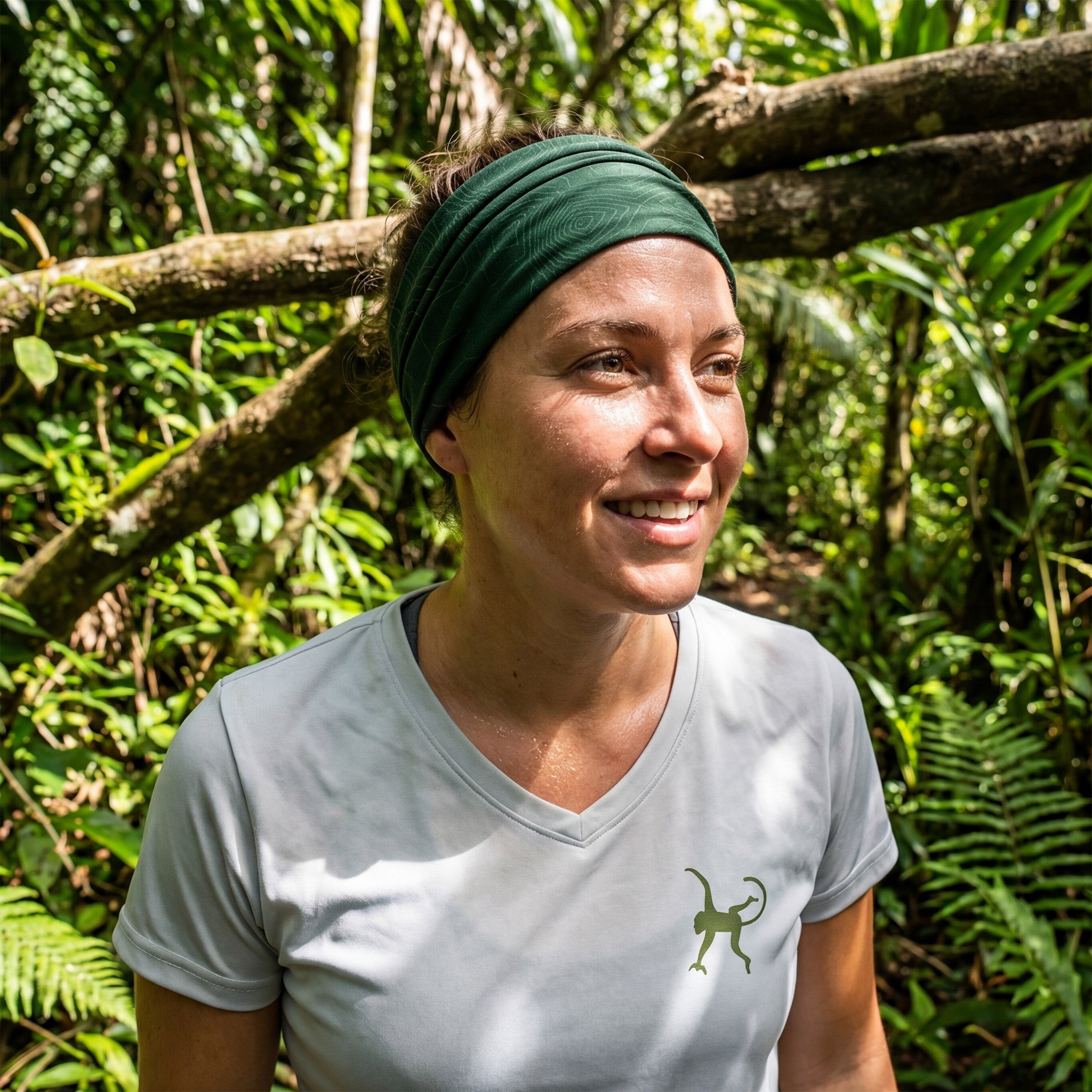 Woman wearing a green neck gaiter as a headband and a light gray UPF shirt with a logo, standing in a forest.