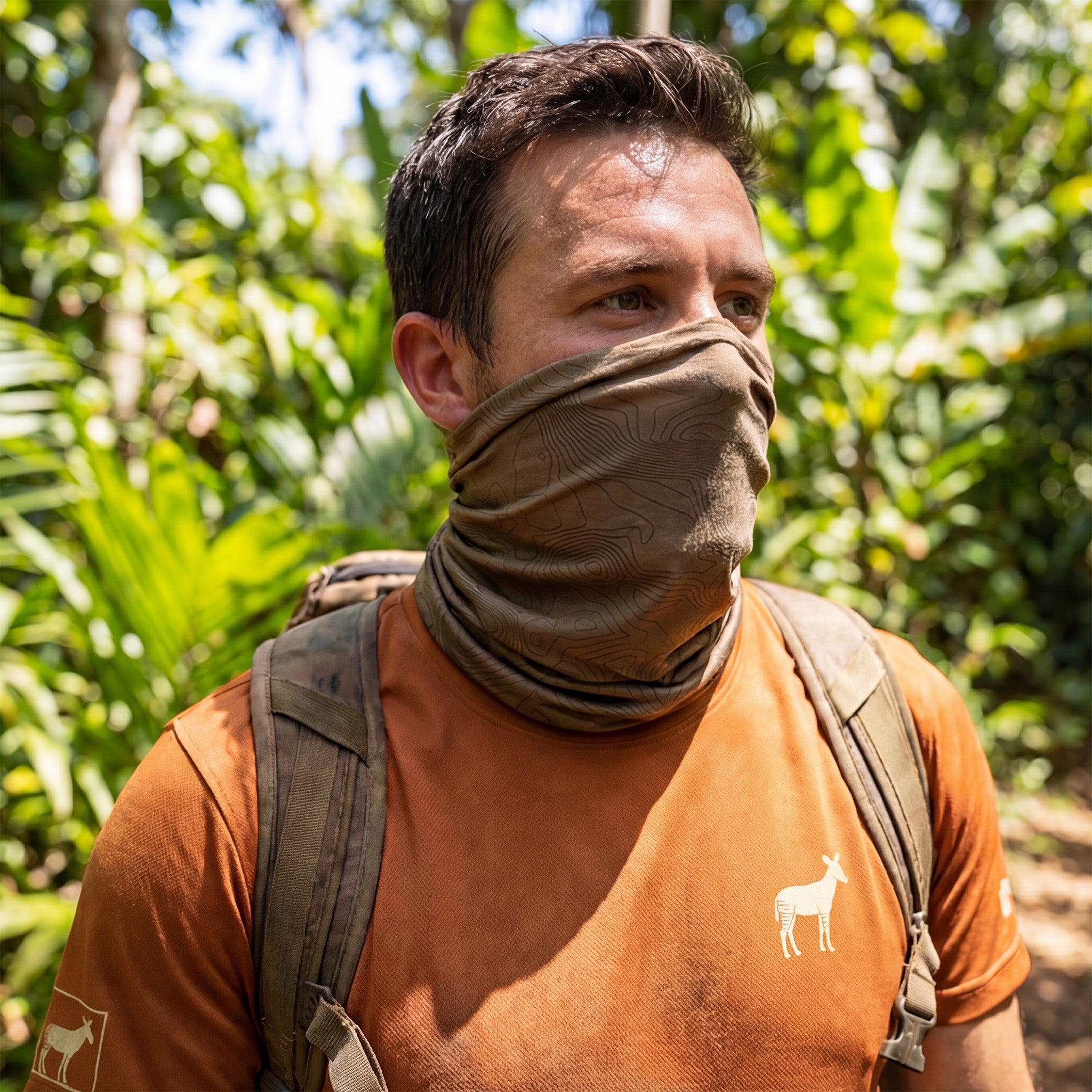 Man wearing a orange shirt with a logo, tan neck gaiter, and backpack in a forest setting