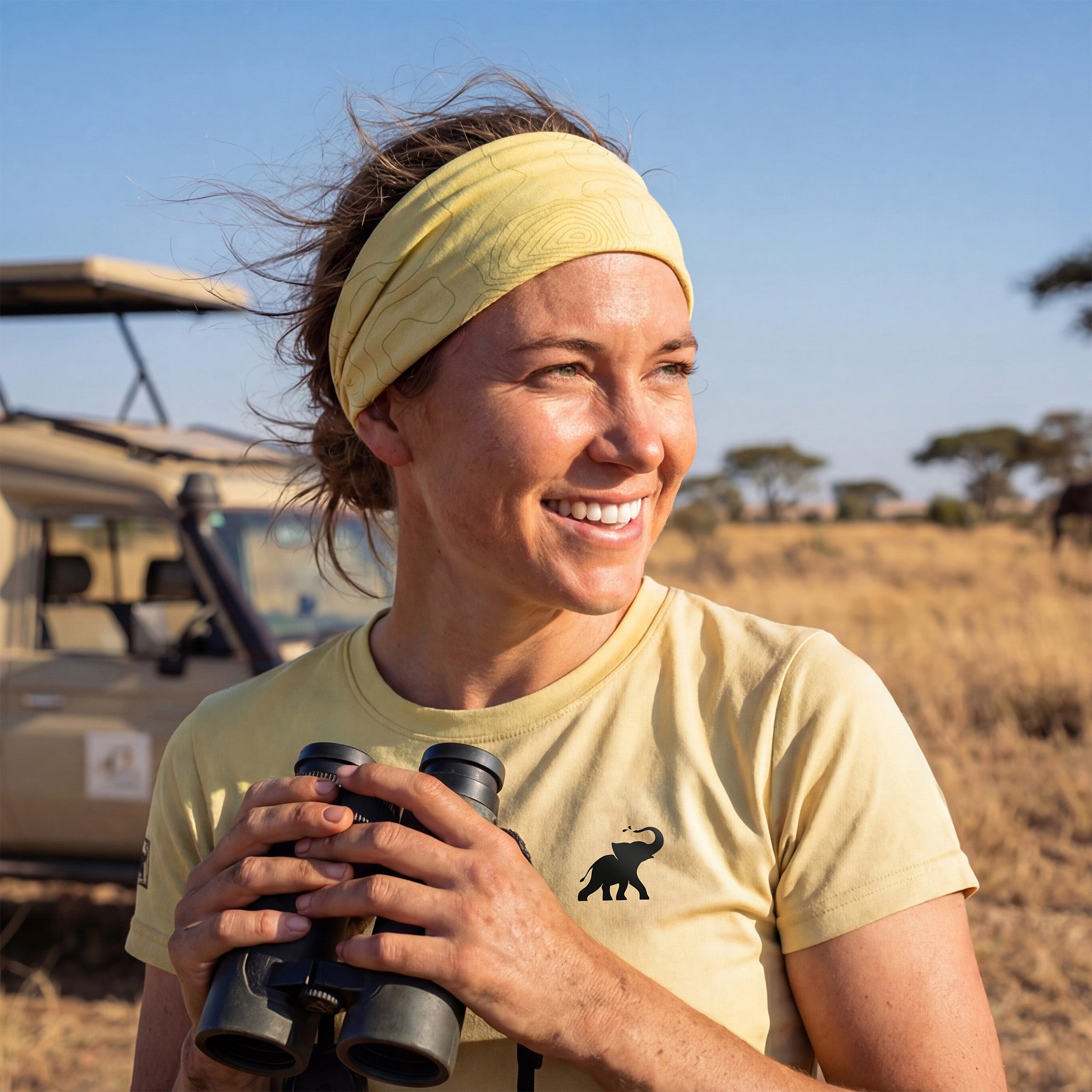 Woman holding binoculars in a safari setting with a vehicle and trees in the background. She's wearing a neck gaiter as a headband and a short sleeve UPF shirt.